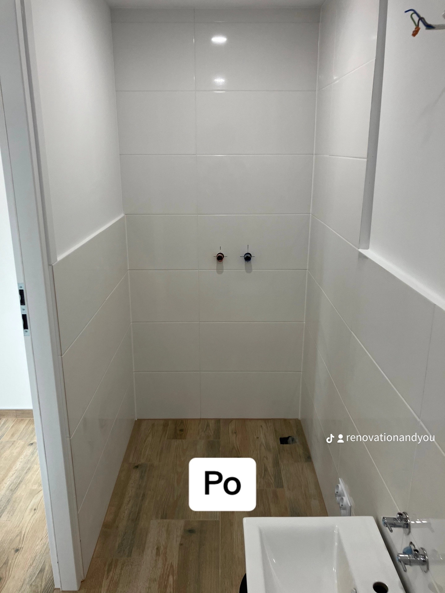 Wnętrze kabiny prysznicowej po remoncie: białe płytki ścienne do połowy wysokości, drewnopodobne płytki podłogowe, widoczne przyłącza wodne i fragment umywalki z baterią.