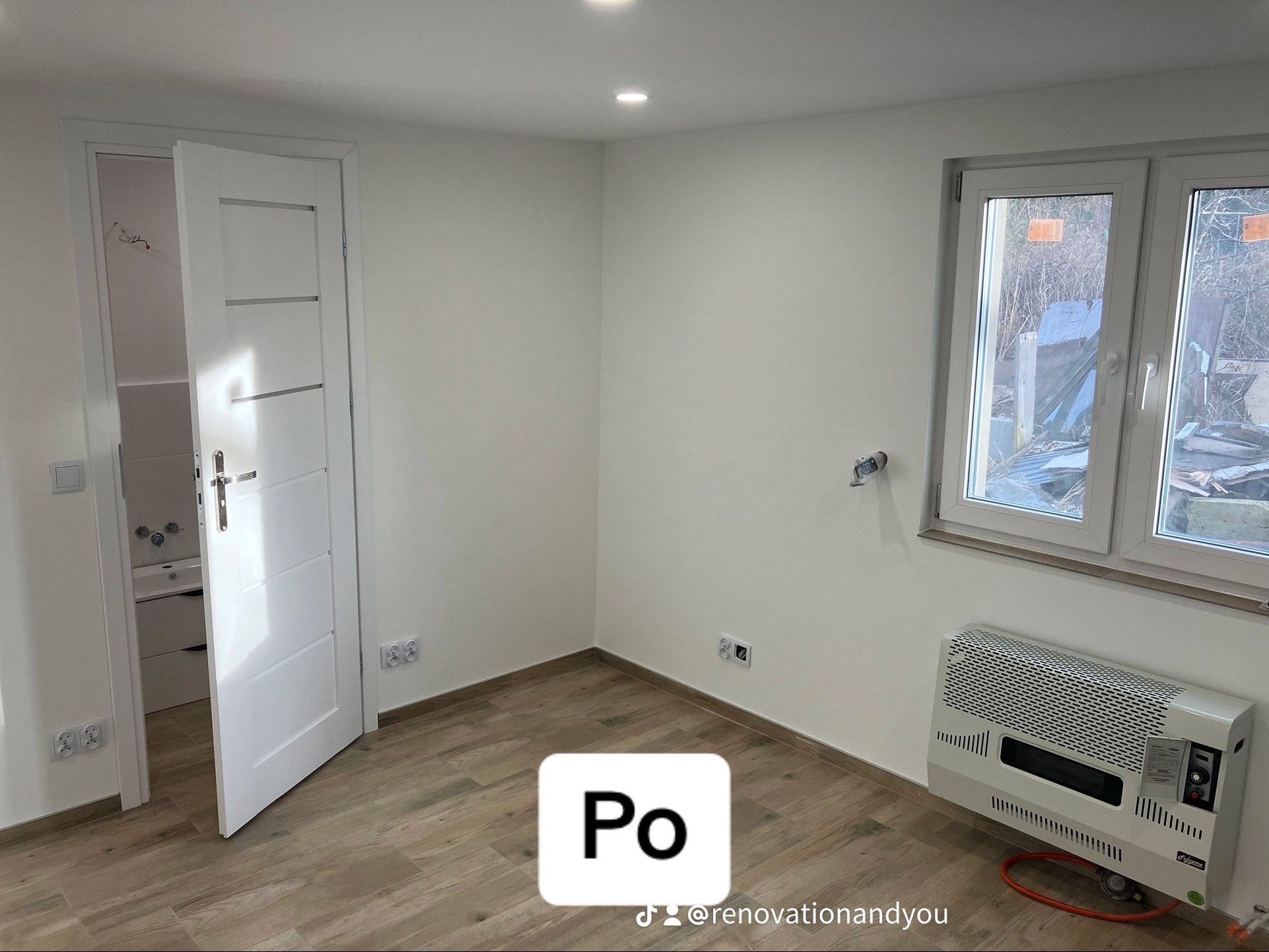 Jasne wnętrze po remoncie: drewnopodobna podłoga, białe ściany z gniazdkami, białe okno z widokiem na zewnątrz i grzejnik gazowy pod nim, otwarte białe drzwi do łazienki z widoczną umywalką.