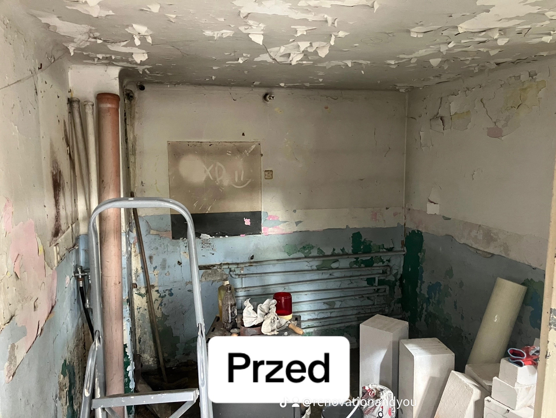 Zdjęcie przedstawia wnętrze pomieszczenia w stanie przed remontem: odpadający tynk na ścianach i suficie, stara instalacja grzewcza, drabina malarska i gruz. Na ścianie widoczne graffiti. Nałożony...
