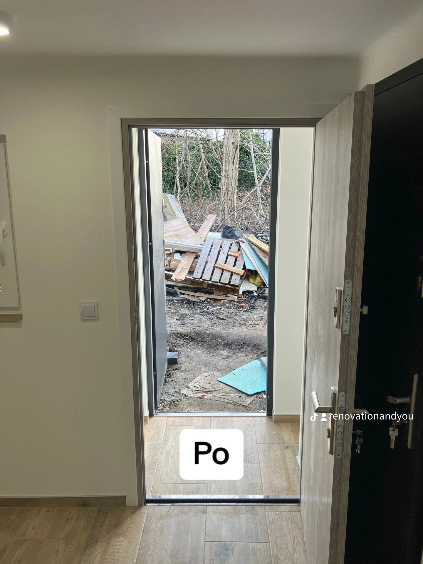 Widok z wnętrza pomieszczenia przez otwarte drzwi na zewnątrz, gdzie widać gruz i materiały budowlane; podłoga wyłożona płytkami imitującymi drewno, na drzwiach widoczny uchwyt i klucze.