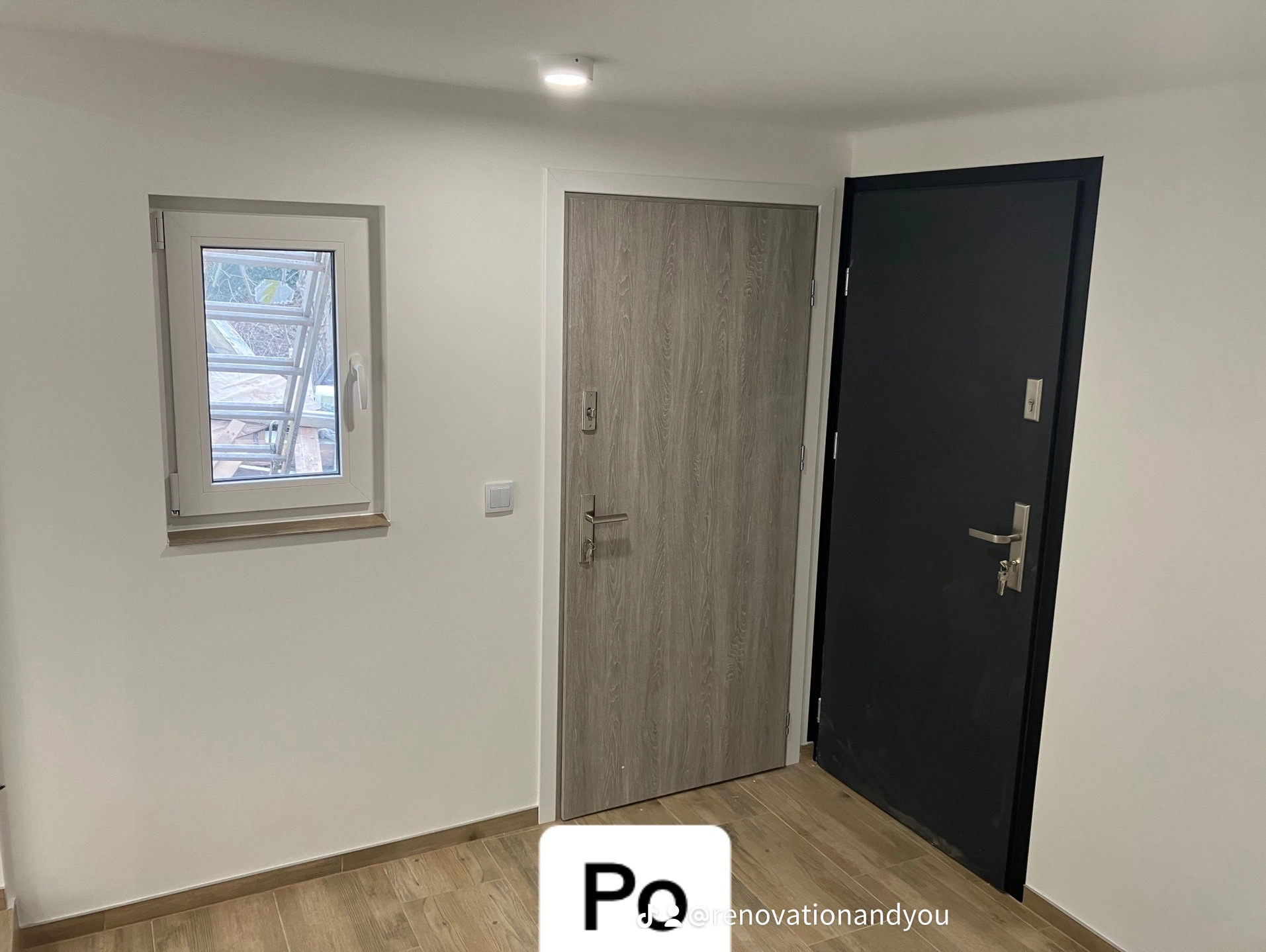 Wnętrze z dwoma drzwiami: drewnopodobne drzwi wejściowe obok czarnych drzwi, małe okno z widokiem na zewnątrz, jasne ściany i podłoga z paneli imitujących drewno.