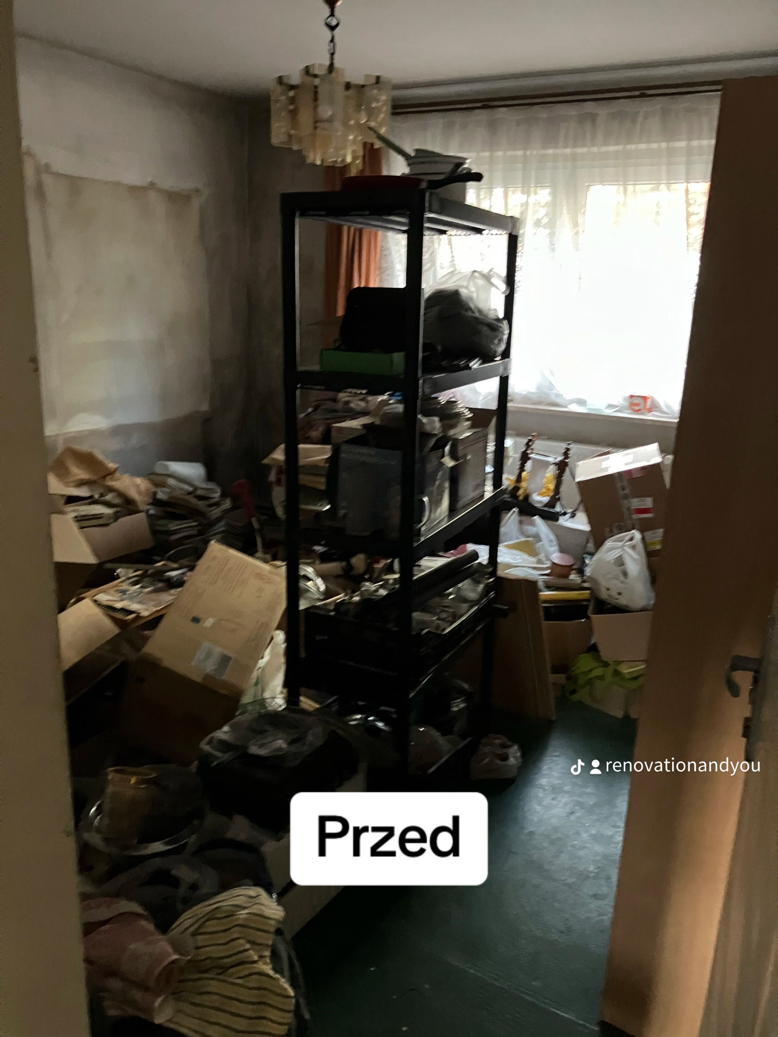 Pokój zagracony kartonami i przedmiotami przed remontem, widoczny regał z półkami, ściany do odświeżenia, okno z zasłonami w tle.
