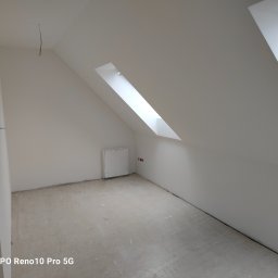 Zakład ogólnobudowlany Jacek oleszczuk - Poddasze ze skośnymi ścianami i oknem dachowym, przygotowane do dalszych prac wykończeniowych, widoczne wystające kable i niezabezpieczone gniazdka elektryczne.