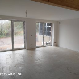 Zakład ogólnobudowlany Jacek oleszczuk - Surowe wnętrze z dwoma oknami balkonowymi, ściany przygotowane do malowania, widoczne wystające kable elektryczne i drewniana belka stropowa.