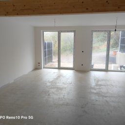 Zakład ogólnobudowlany Jacek oleszczuk - Surowe, jasne wnętrze z betonową podłogą przed wykończeniem, widoczne okna z widokiem na zewnątrz i surowe drewniane belki stropowe.