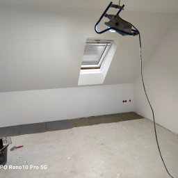 Zakład ogólnobudowlany Jacek oleszczuk - Poddasze w trakcie układania szarych płytek podłogowych, widoczny kubeł z zaprawą, poziomica i szpachelka, oświetlenie robocze, okno dachowe.