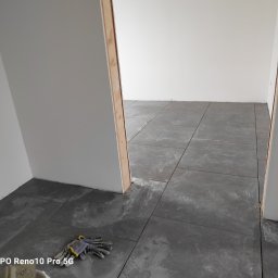 Zakład ogólnobudowlany Jacek oleszczuk - Szare płytki podłogowe ułożone w pokoju, widoczne robocze rękawice w rogu, surowe ściany i ościeżnica.