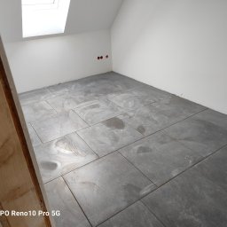 Zakład ogólnobudowlany Jacek oleszczuk - Świeżo ułożone, szare płytki podłogowe w pomieszczeniu na poddaszu z oknem dachowym, widoczne ślady po czyszczeniu, fragment drewnianej konstrukcji w lewym dolnym rogu.