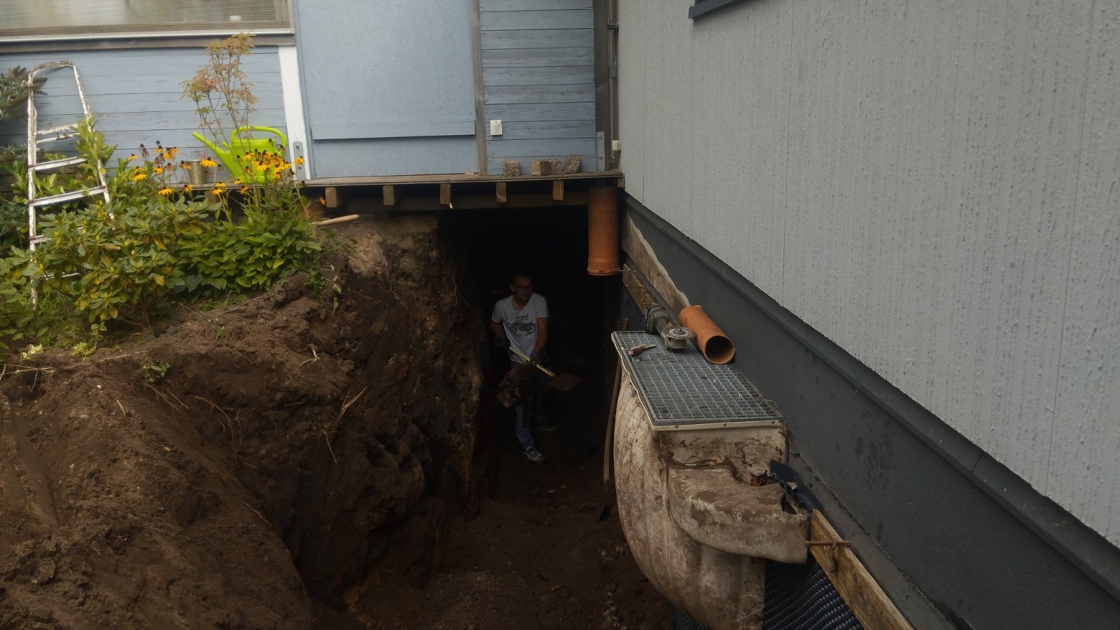 Wykop fundamentów w ziemi przy budynku, widoczny pracownik z łopatą w wykopie, fragment instalacji kanalizacyjnej z kratką i rurami.