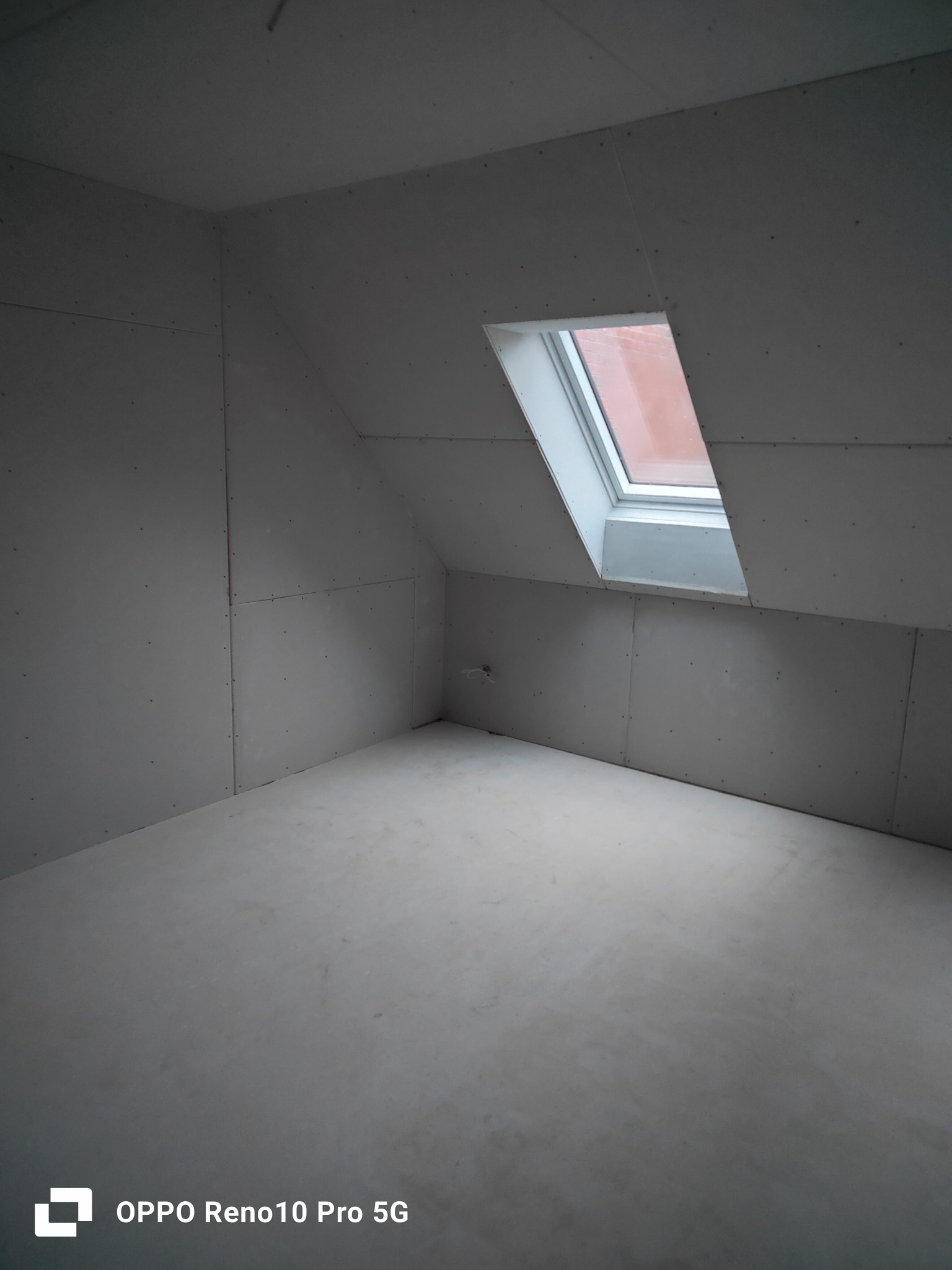 Pokój na poddaszu z oknem dachowym, ściany wyłożone płytami gipsowo-kartonowymi, widoczne miejsca łączeń i wkręty, podłoga przygotowana do dalszych prac wykończeniowych.