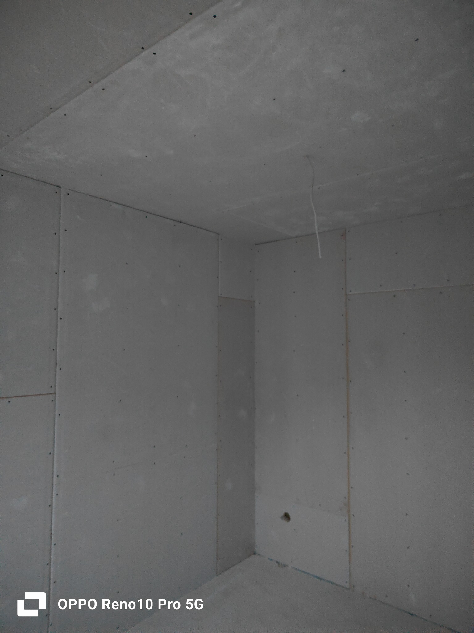 Surowe wnętrze z zamontowanymi płytami gipsowo-kartonowymi na ścianach i suficie, widoczne łączenia i śruby, pojedynczy kabel zwisający z sufitu.