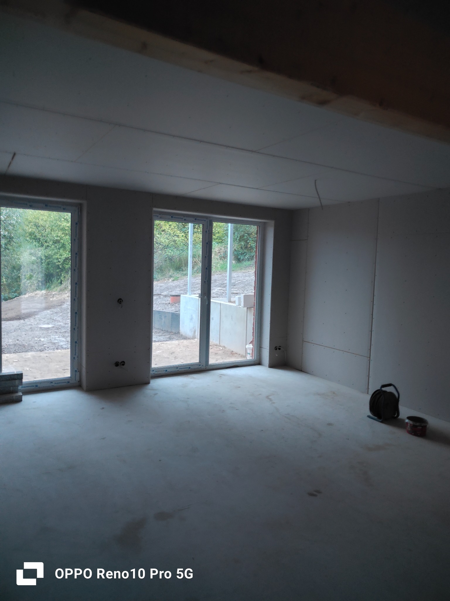 Wnętrze w trakcie prac wykończeniowych: ściany obłożone płytami, widoczne okna z widokiem na zewnątrz, na podłodze leżą narzędzia i puszka farby.