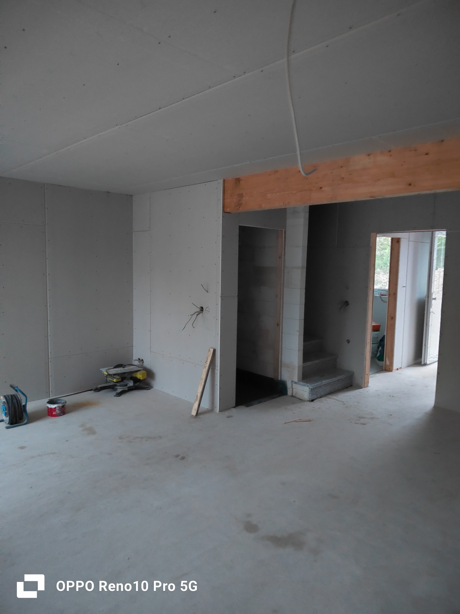 Wnętrze w trakcie montażu ścian z płyt, widoczne narzędzia, drewniana belka nad przejściem do innego pomieszczenia i częściowo wykończone schody.