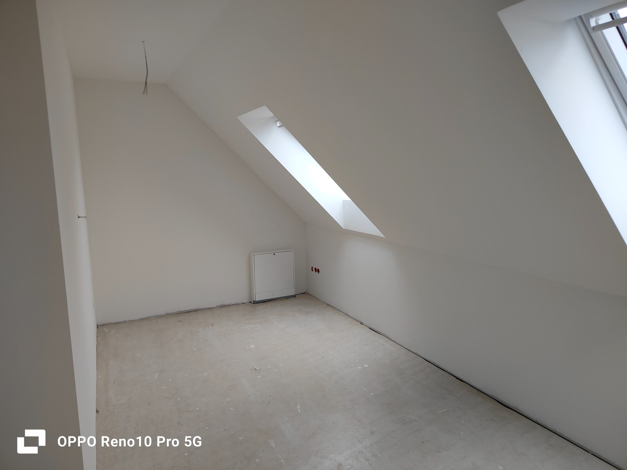 Poddasze ze skośnymi ścianami i oknem dachowym, przygotowane do dalszych prac wykończeniowych, widoczne wystające kable i niezabezpieczone gniazdka elektryczne.