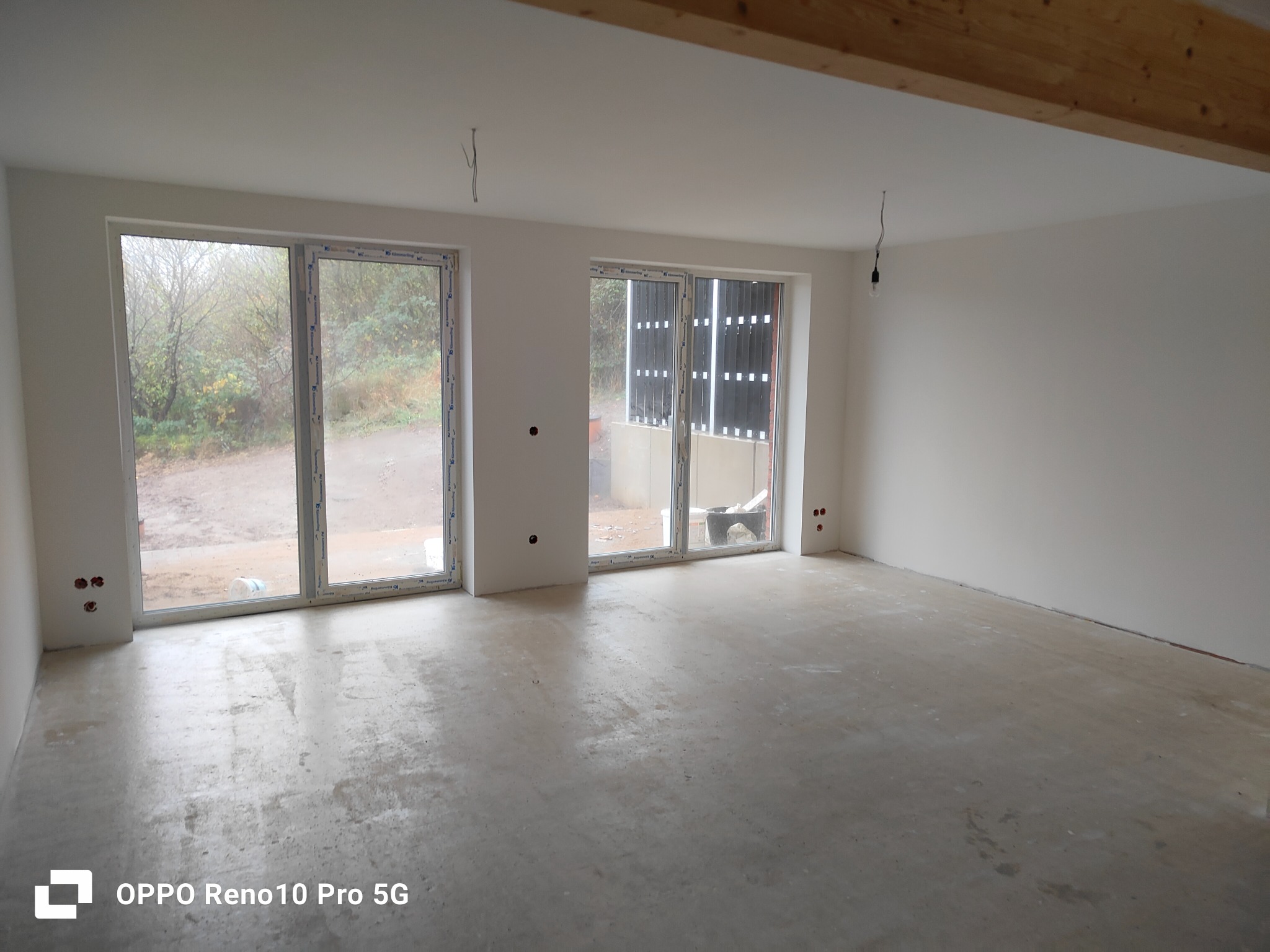 Surowe wnętrze z dwoma oknami balkonowymi, ściany przygotowane do malowania, widoczne wystające kable elektryczne i drewniana belka stropowa.