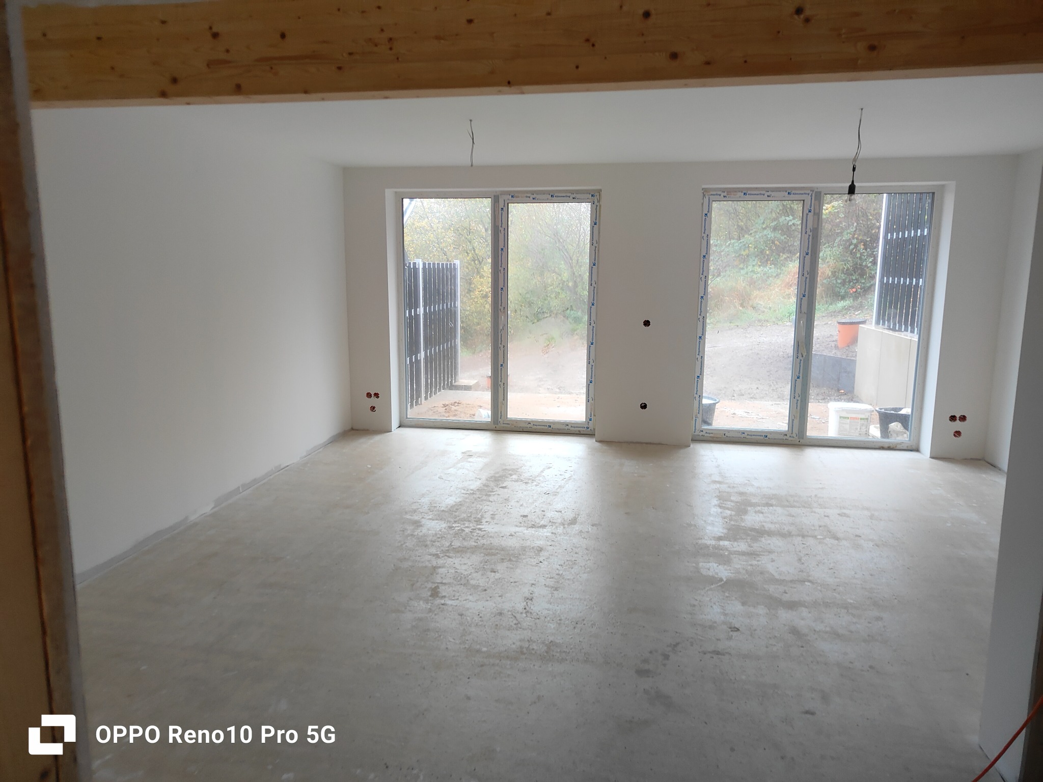 Surowe, jasne wnętrze z betonową podłogą przed wykończeniem, widoczne okna z widokiem na zewnątrz i surowe drewniane belki stropowe.