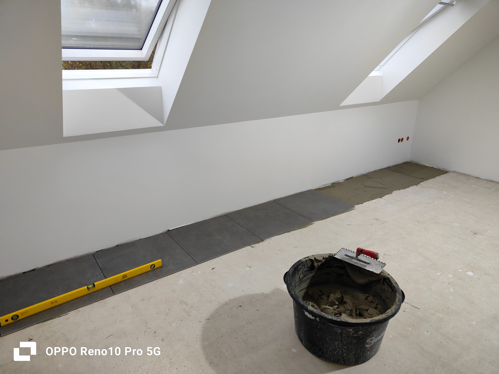 Przygotowanie do układania płytek podłogowych na poddaszu, widoczny poziomica, wiadro z zaprawą klejową i szpachelka, część płytek już ułożona.