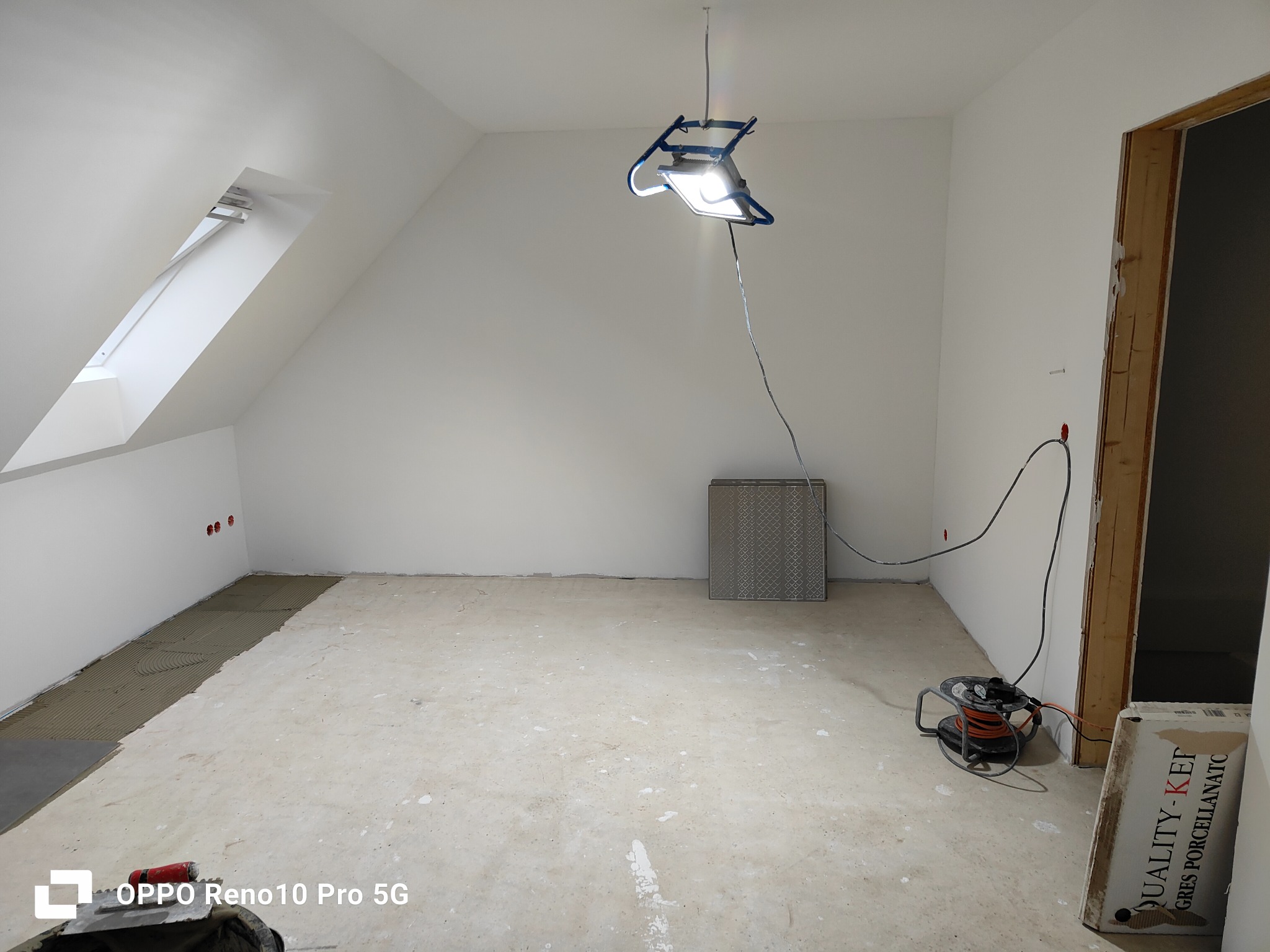 Poddasze w trakcie prac: częściowo ułożone płytki podłogowe, widoczny klej, lampa budowlana, zwinięty przedłużacz oraz karton z płytkami podłogowymi Quality-Kef Gres Porcellanato.