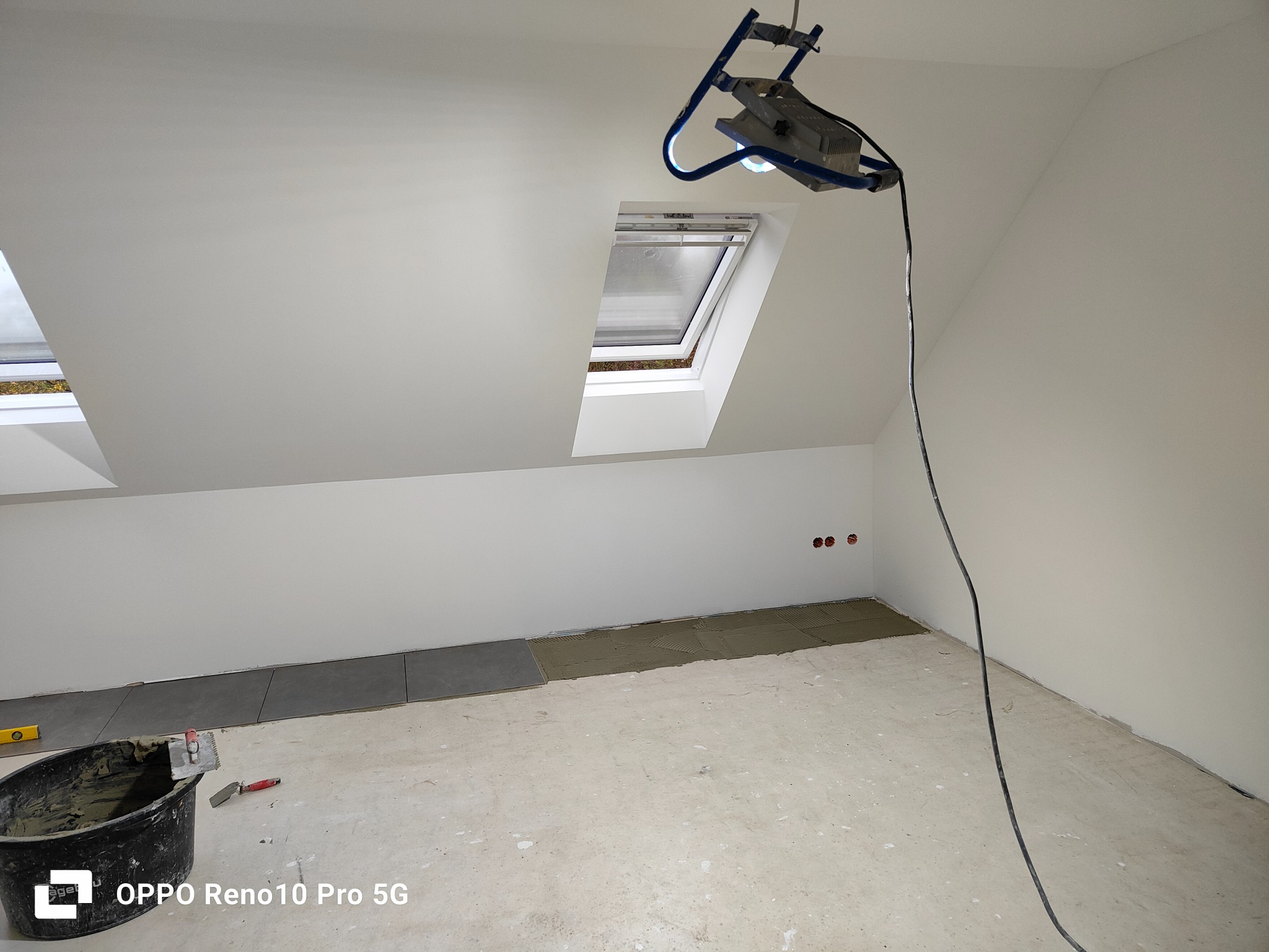 Poddasze w trakcie układania szarych płytek podłogowych, widoczny kubeł z zaprawą, poziomica i szpachelka, oświetlenie robocze, okno dachowe.