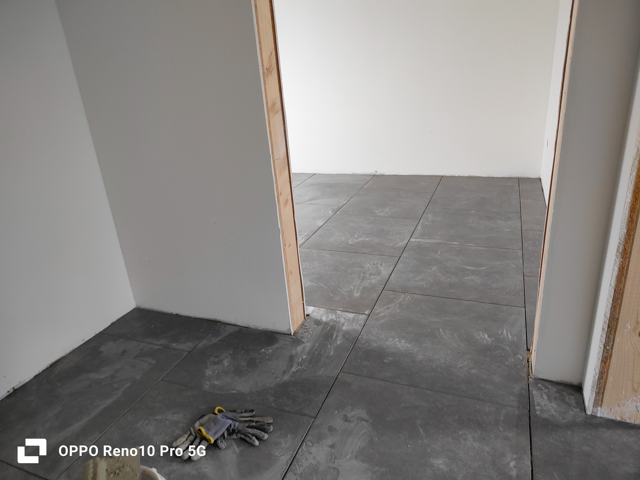 Szare płytki podłogowe ułożone w pokoju, widoczne robocze rękawice w rogu, surowe ściany i ościeżnica.