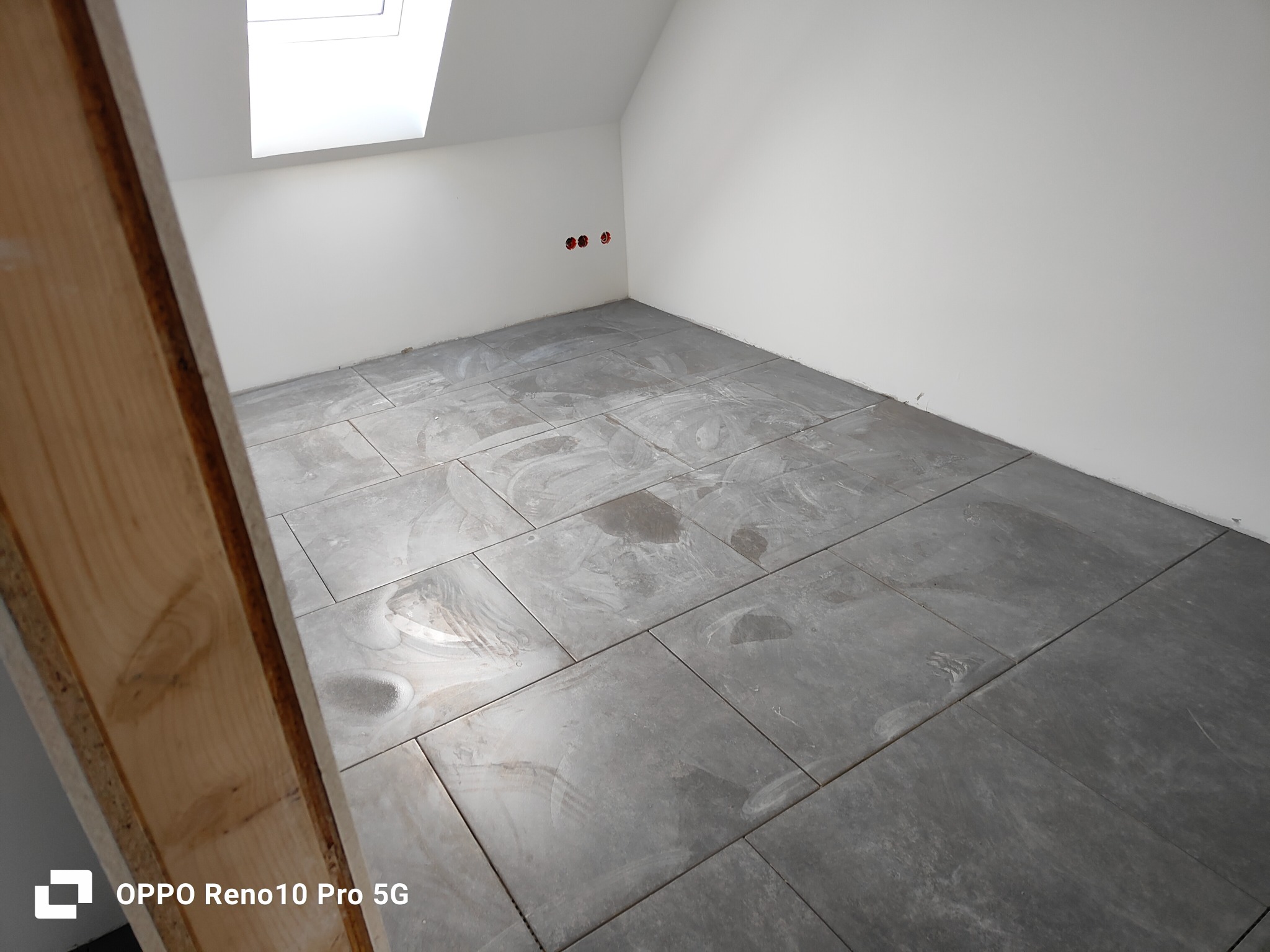 Świeżo ułożone, szare płytki podłogowe w pomieszczeniu na poddaszu z oknem dachowym, widoczne ślady po czyszczeniu, fragment drewnianej konstrukcji w lewym dolnym rogu.