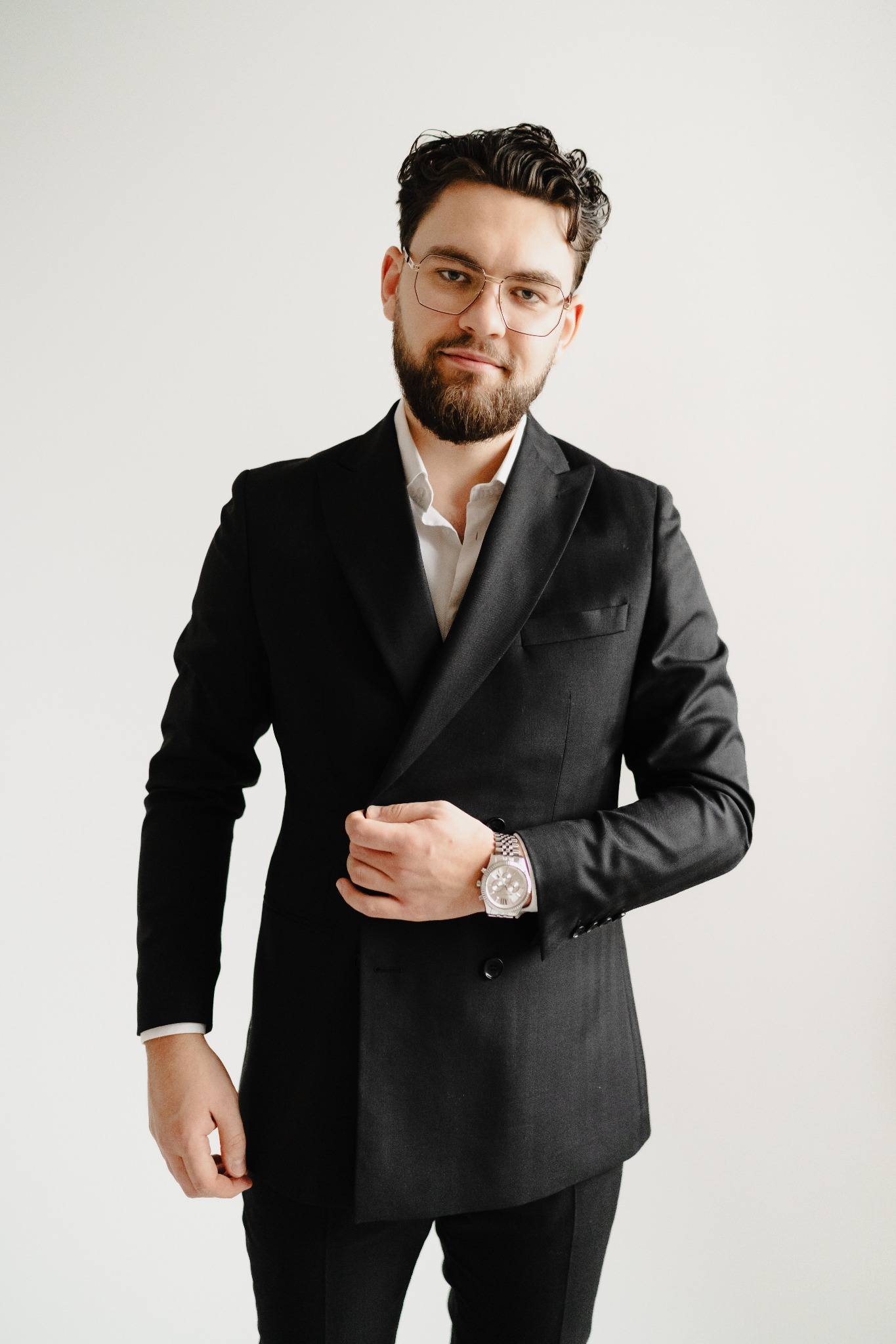 Portret mężczyzny w czarnym garniturze, białej koszuli i okularach w złotych oprawkach, z brodą i zegarkiem na ręce, na jasnym tle.