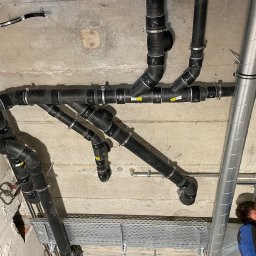 Radosław Korneusz - Skomplikowana instalacja rur kanalizacyjnych i wentylacyjnych pod betonowym sufitem, widoczne czarne rury z białymi oznaczeniami 'X', metalowe obejmy, koryto kablowe oraz fragment sylwetki...