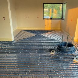 Radosław Korneusz - Montaż ogrzewania podłogowego w surowym wnętrzu, widoczne rury grzewcze ułożone na folii z siatką, zwinięty zapas rur, surowy betonowy sufit i okno z widokiem na zieleń.