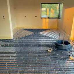 Montaż ogrzewania podłogowego w surowym wnętrzu, widoczne rury grzewcze ułożone na folii z siatką, zwinięty zapas rur, surowy betonowy sufit i okno z widokiem na zieleń.