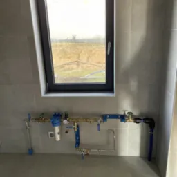 Kompletna instalacja wodna z filtrem, manometrem i rurami miedzianymi oraz niebieskimi elementami, zamontowana na szarej ścianie z płytkami, z oknem w tle.