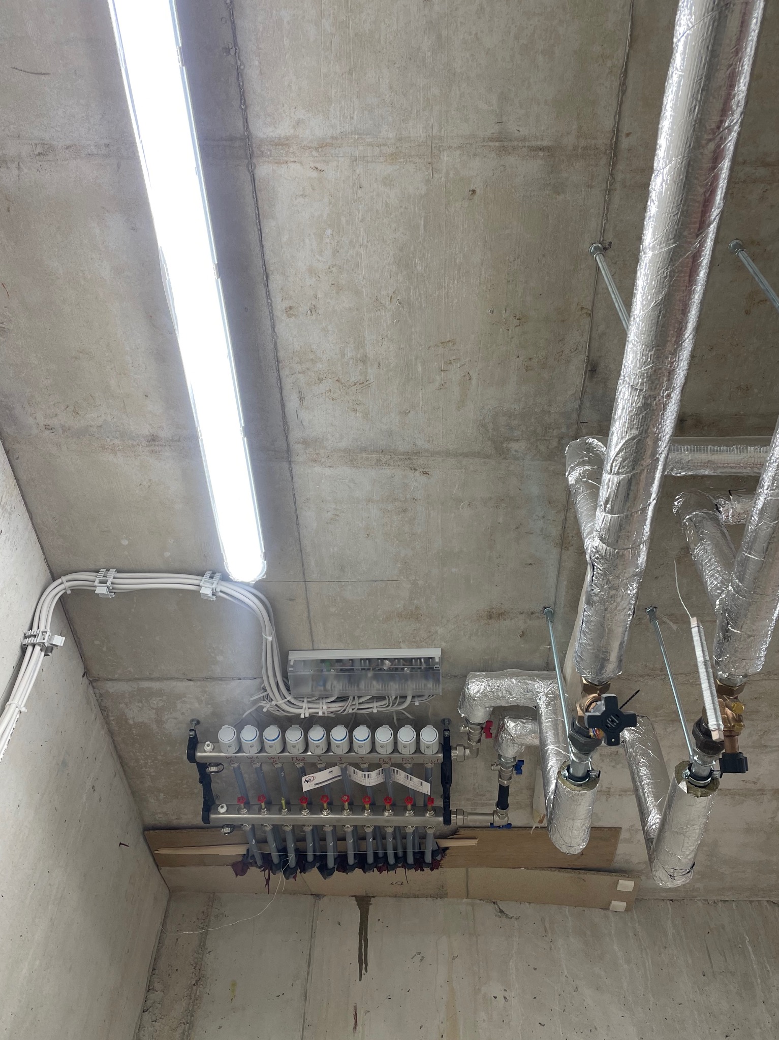 Rozdzielacz ogrzewania podłogowego z rurami w izolacji aluminiowej, zamontowany na betonowym suficie z widocznymi przewodami elektrycznymi i oświetleniem jarzeniowym.