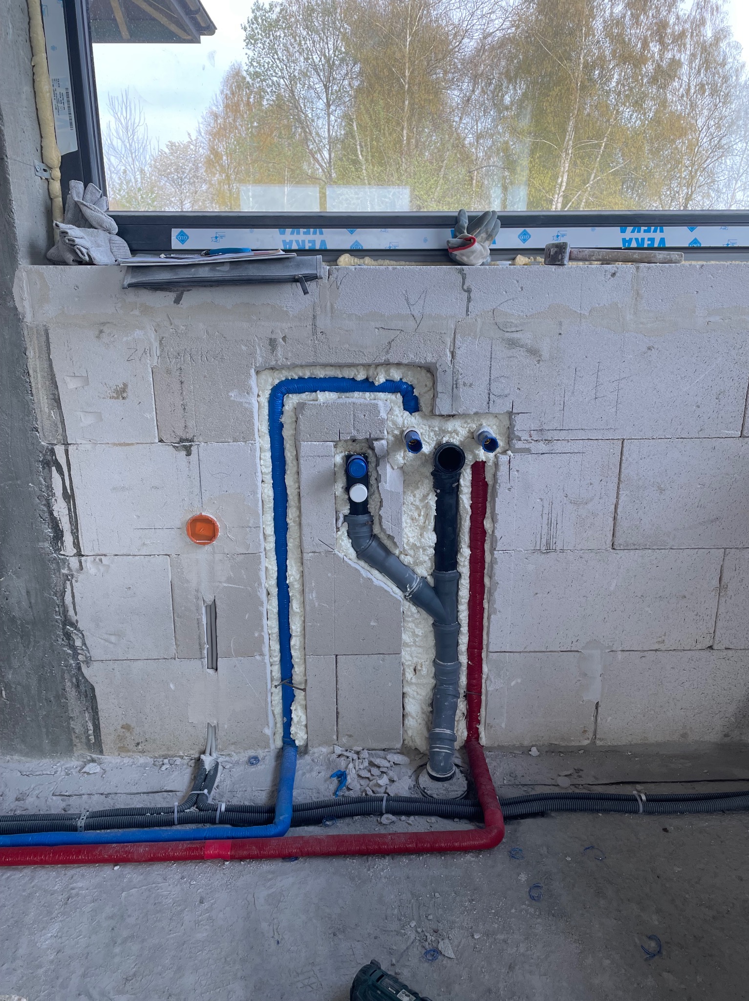Wnęka w ścianie z betonu komórkowego, w której umieszczono instalację wodno-kanalizacyjną z rur w kolorach niebieskim i czerwonym, zabezpieczoną pianką montażową. Widoczne rury kanalizacyjne...
