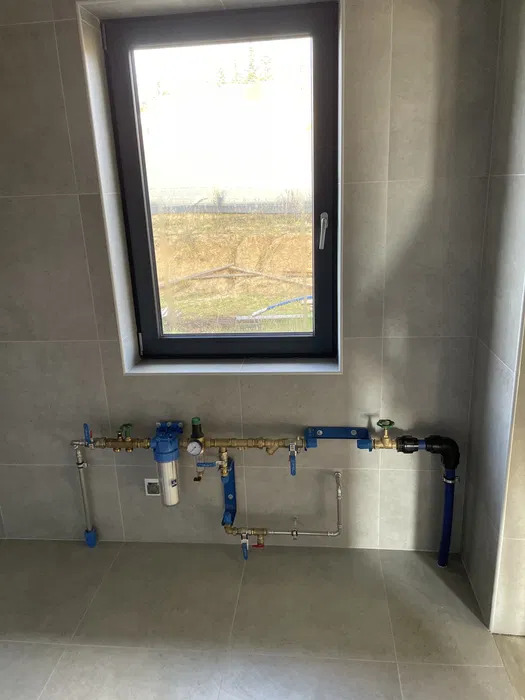 Kompletna instalacja wodna z filtrem, manometrem i rurami miedzianymi oraz niebieskimi elementami, zamontowana na szarej ścianie z płytkami, z oknem w tle.