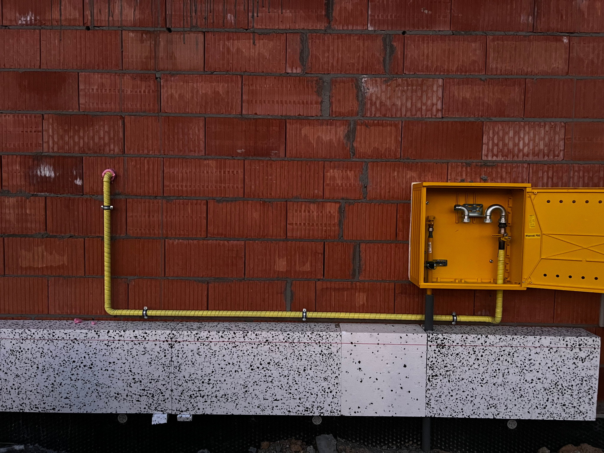 Żółta rura gazowa przymocowana do ściany z czerwonej cegły i białego styropianu, z otwartą żółtą skrzynką gazową po prawej stronie.