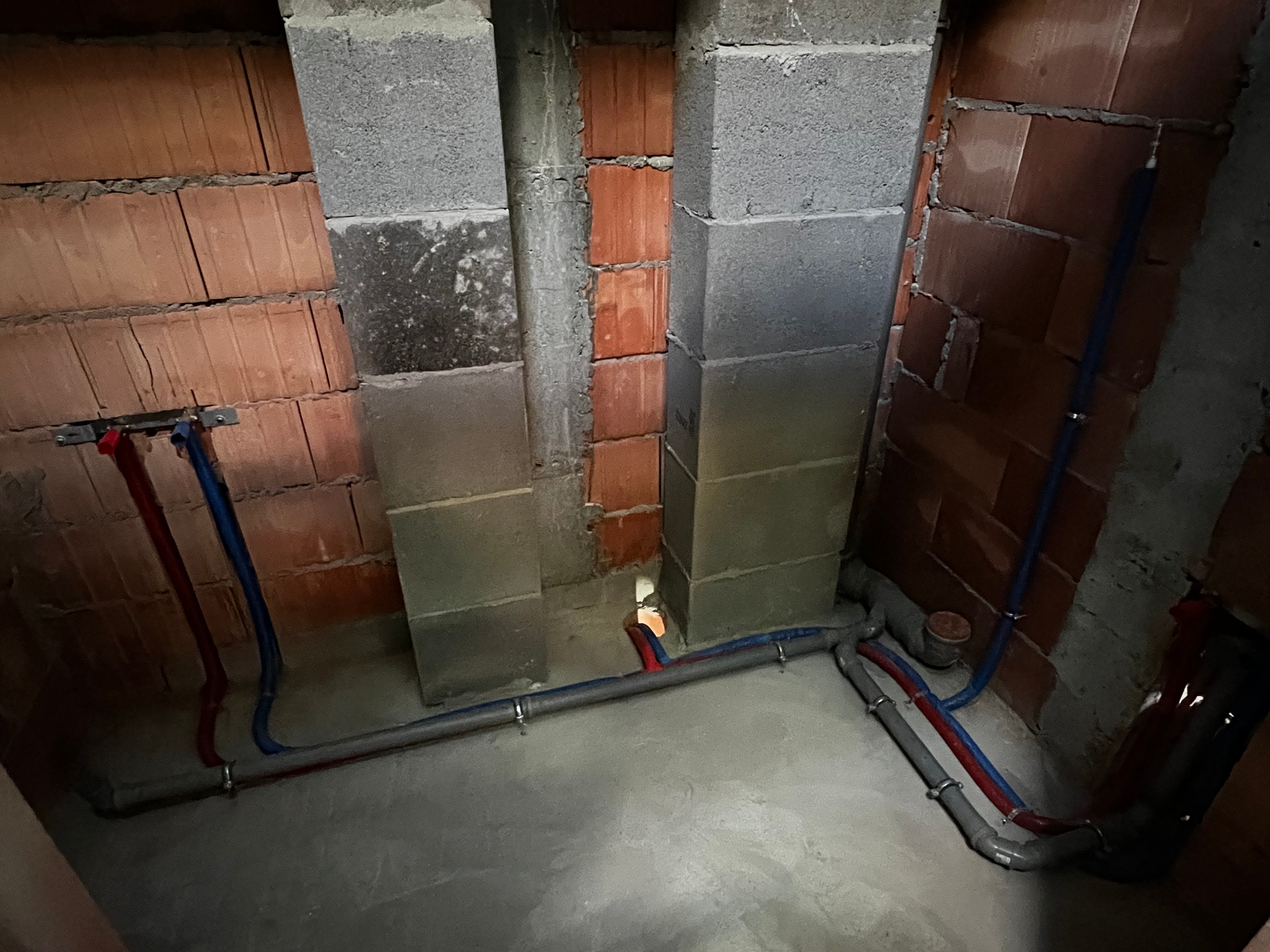 Surowy stan instalacji hydraulicznej w narożniku pomieszczenia z ceglanymi i betonowymi ścianami, widoczne rury w kolorach niebieskim i czerwonym mocowane do podłoża.