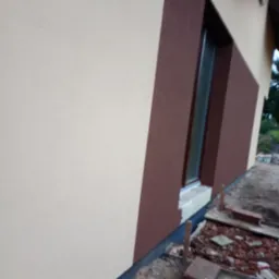 Fragment elewacji domu po nałożeniu tynku w dwóch odcieniach beżu i brązu, widoczne okno z parapetem, teren wokół domu w trakcie prac wykończeniowych.