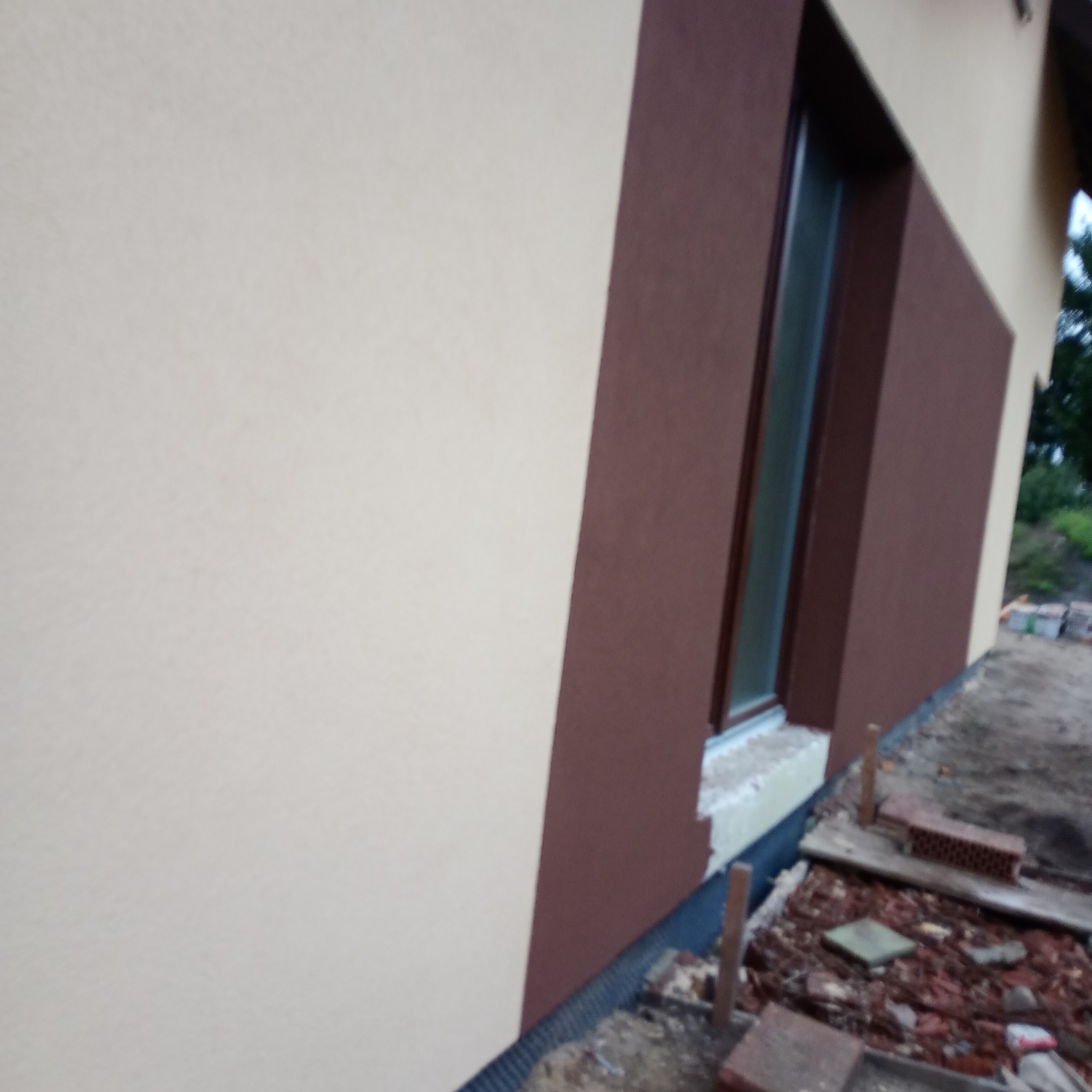 Fragment elewacji domu po nałożeniu tynku w dwóch odcieniach beżu i brązu, widoczne okno z parapetem, teren wokół domu w trakcie prac wykończeniowych.