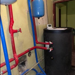 GASP USŁUGI HYDRAULICZNE - Czarny zasobnik ciepłej wody użytkowej z podłączonymi rurami w izolacji czerwonej i niebieskiej, widoczne również manometry i zawory, na górze zbiornika leży biały termostat.