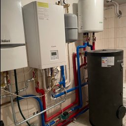 GASP USŁUGI HYDRAULICZNE - Kotłownia z dwoma piecami gazowymi, zasobnikiem ciepłej wody użytkowej Cubion, naczyniami wzbiorczymi i instalacją hydrauliczną z rur w kolorach czerwonym i niebieskim na tle beżowych płytek.