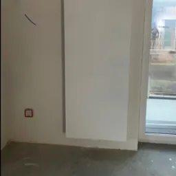 Biały, panelowy grzejnik zamontowany na ścianie w surowym pomieszczeniu, widoczny wystający kabel i czujnik dymu, okno balkonowe z widokiem na zewnątrz.