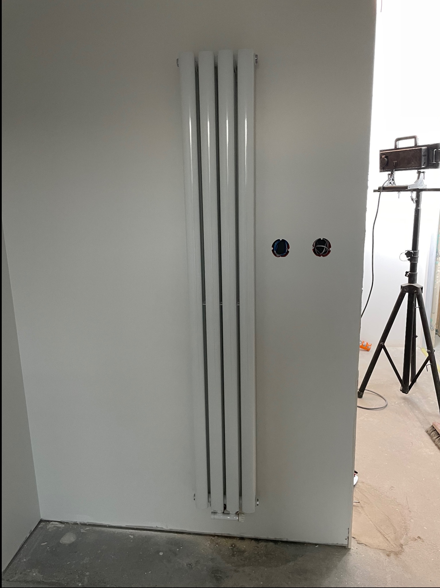 Nowy, pionowy grzejnik panelowy zamontowany na białej ścianie, widoczne puszki elektryczne i lampa studyjna w tle.