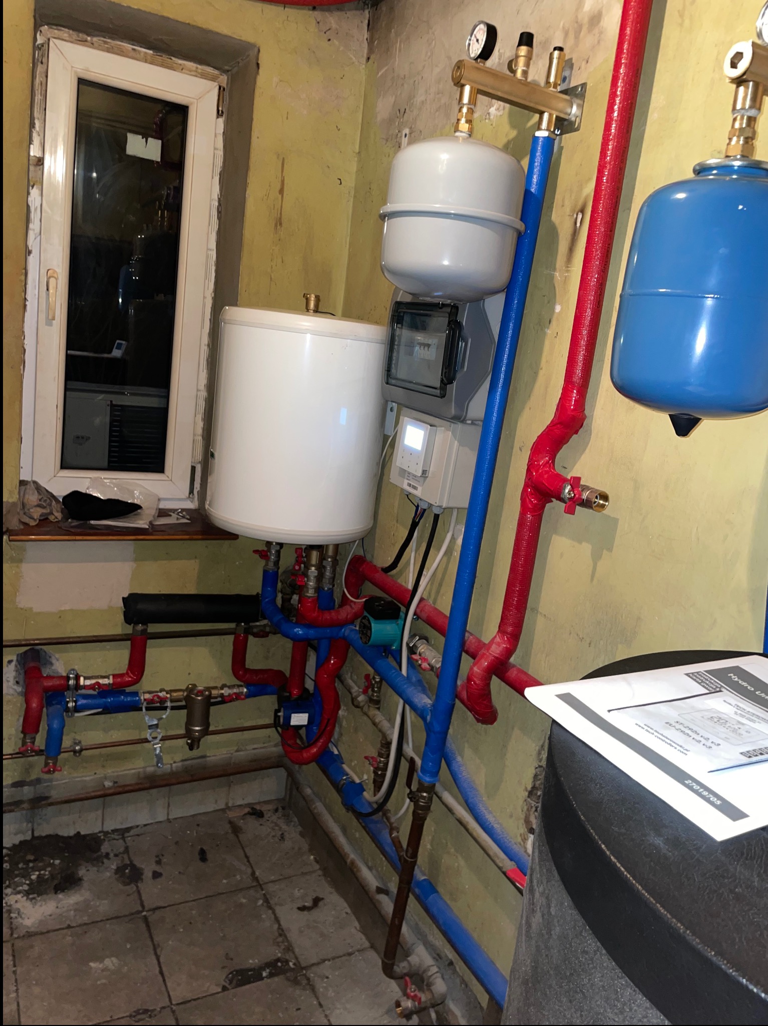 Instalacja centralnego ogrzewania w piwnicy: biały bojler, niebieski zbiornik wyrównawczy, czerwone i niebieskie rury, manometry, sterownik, widoczne przyłącza i zawory, obok okno piwniczne.