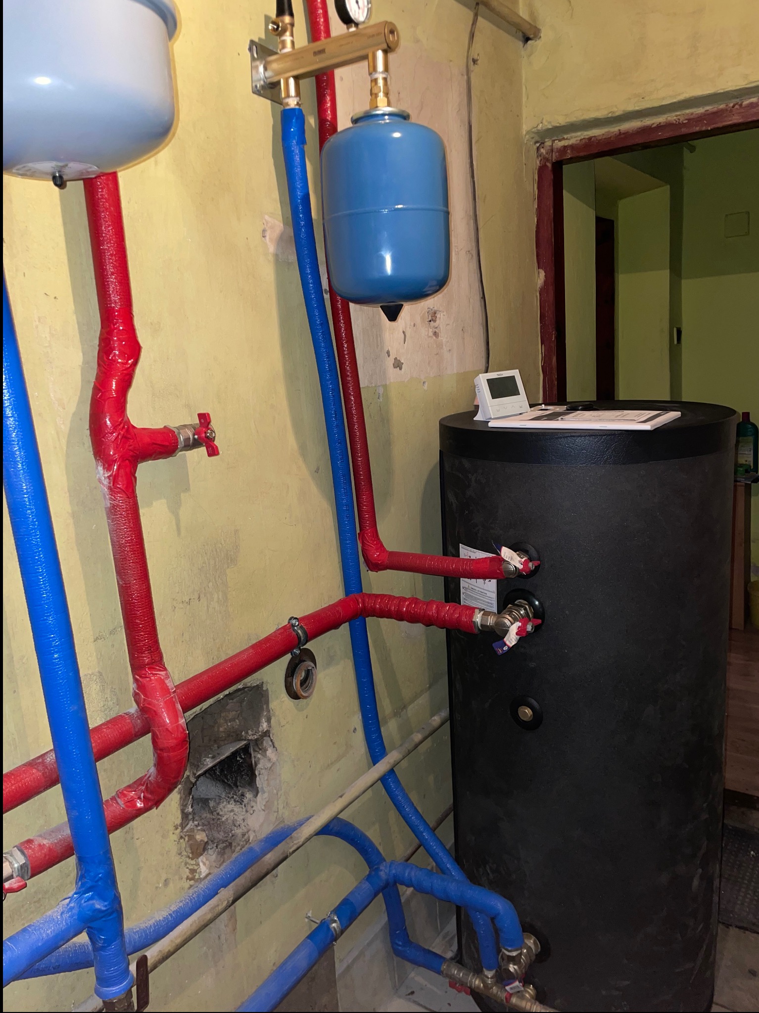 Czarny zasobnik ciepłej wody użytkowej z podłączonymi rurami w izolacji czerwonej i niebieskiej, widoczne również manometry i zawory, na górze zbiornika leży biały termostat.
