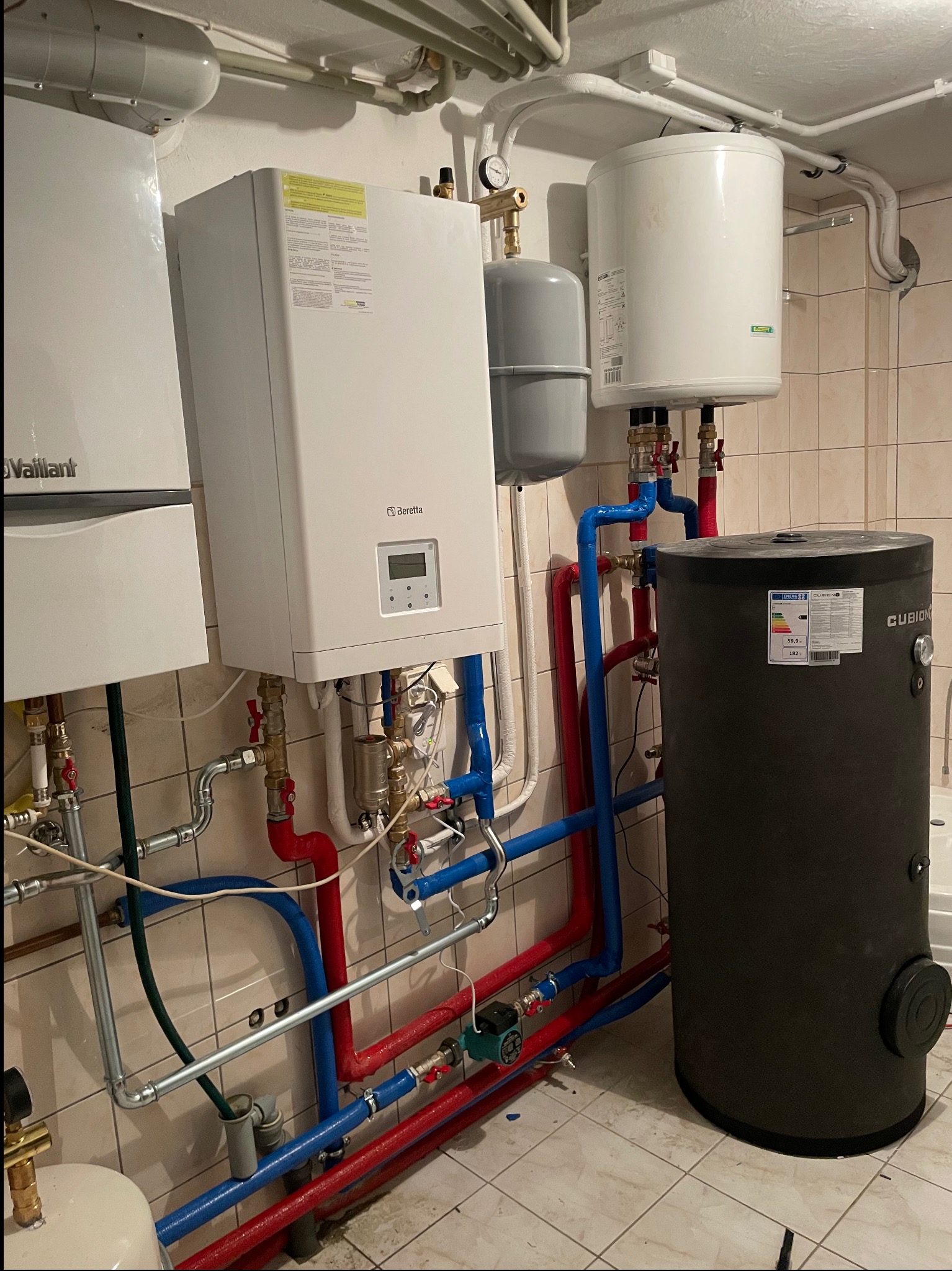 Kotłownia z dwoma piecami gazowymi, zasobnikiem ciepłej wody użytkowej Cubion, naczyniami wzbiorczymi i instalacją hydrauliczną z rur w kolorach czerwonym i niebieskim na tle beżowych płytek.