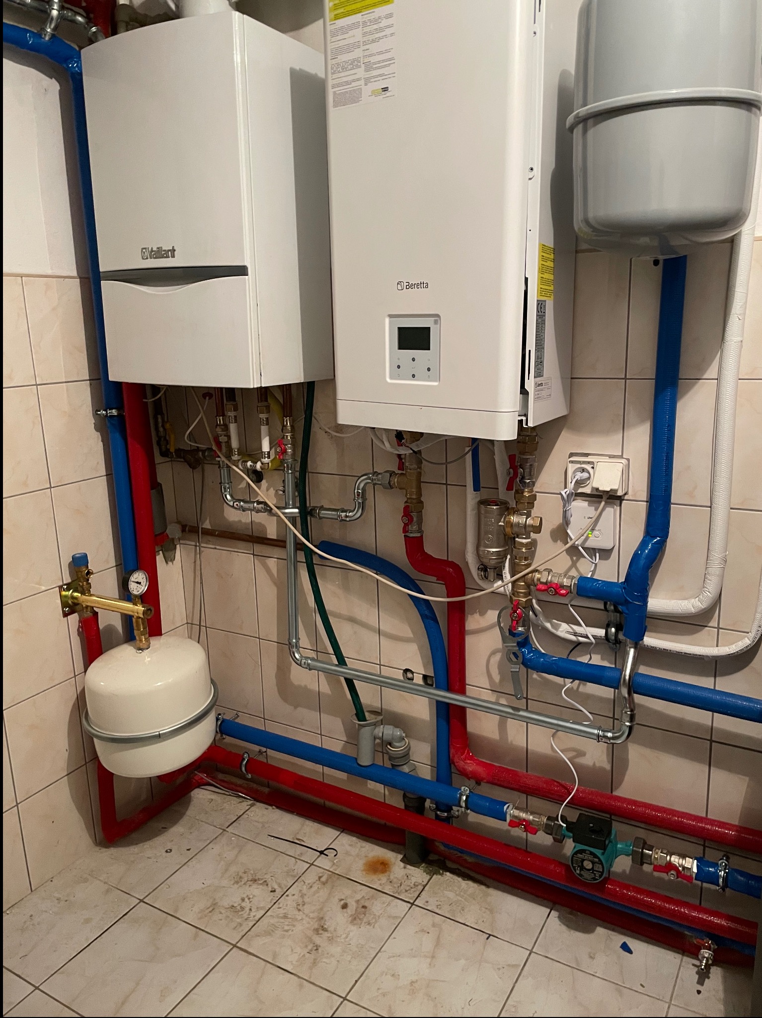Dwa wiszące piece gazowe, Vaillant i Beretta, z podłączeniami rur w kolorach niebieskim i czerwonym, zamontowane na ścianie z płytek w pomieszczeniu gospodarczym.