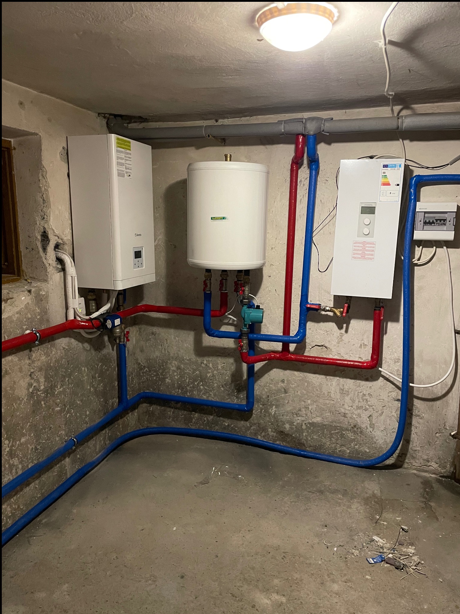 Piwnica z zainstalowanymi dwoma piecami gazowymi marki Beretta i zasobnikiem wody, połączonymi rurami w kolorze czerwonym i niebieskim, pod sufitem widoczna instalacja oraz oświetlenie.