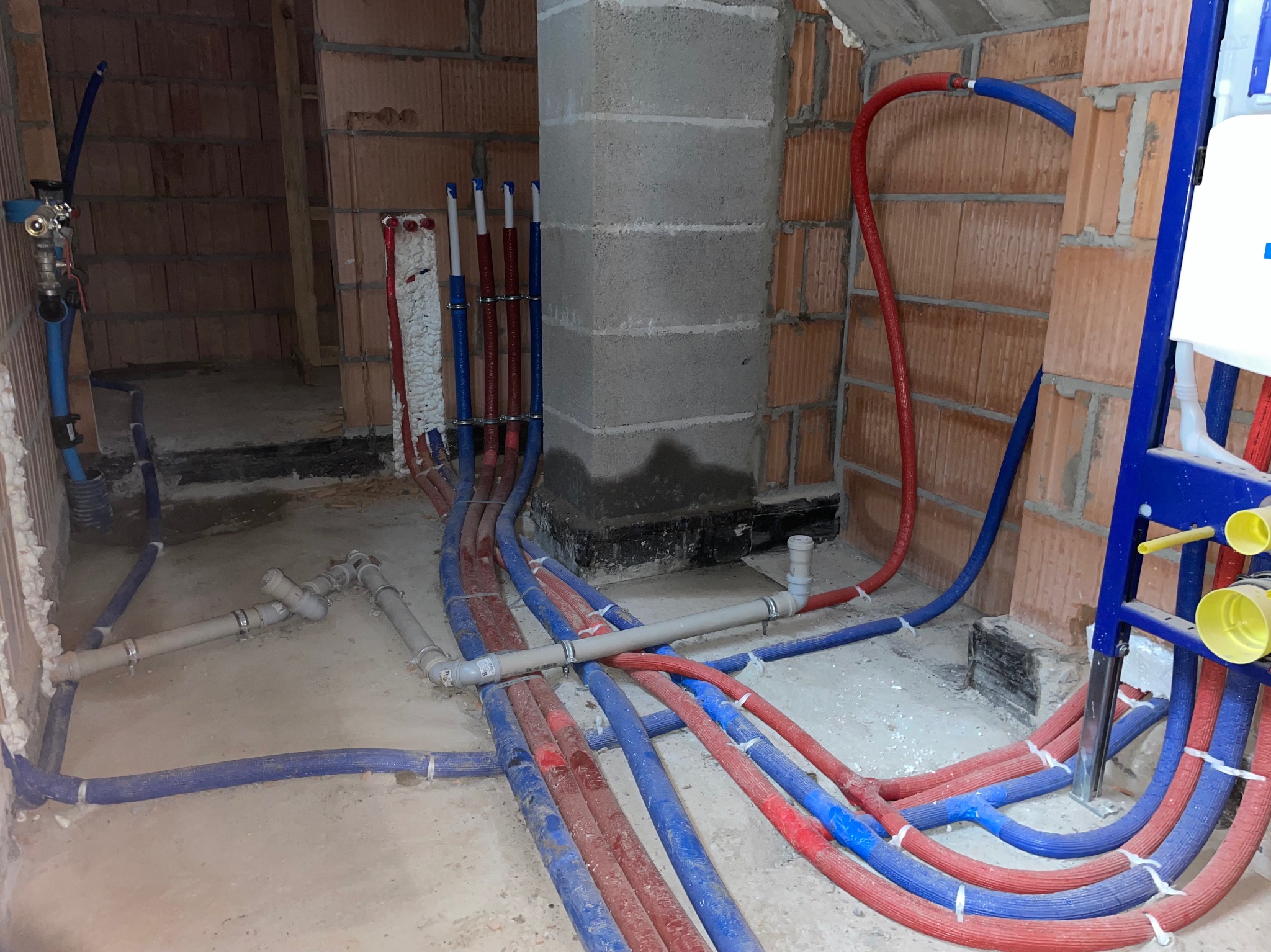 Wielobarwne rury instalacji wodnej i grzewczej w surowym stanie, prowadzone wzdłuż ściany z czerwonej cegły i betonowego filaru, z widocznym stelażem podtynkowym WC.