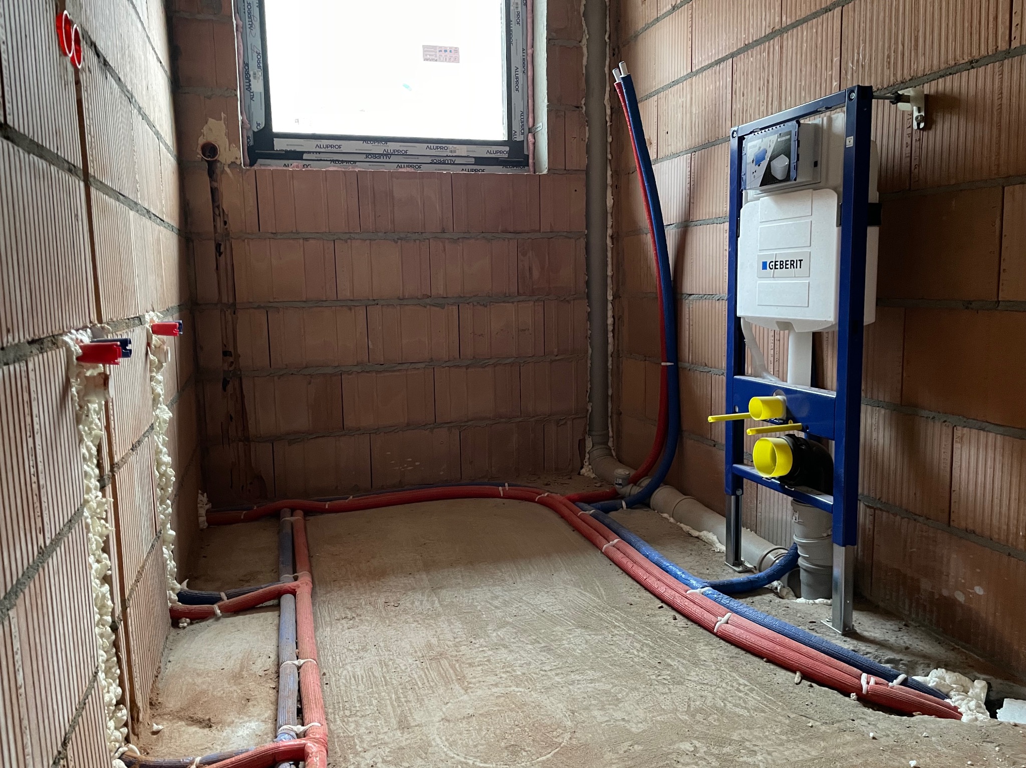 Surowy stan łazienki w trakcie instalacji. Widoczne czerwono-niebieskie rury rozprowadzone po podłodze i ścianach, zamocowany stelaż podtynkowy Geberit z żółtymi elementami, okno w stanie surowym...
