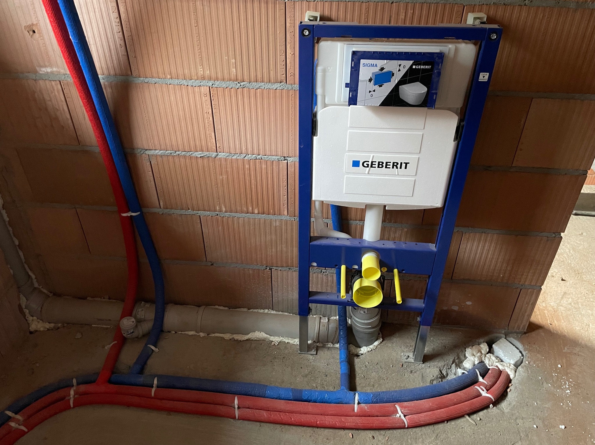 Stelaż podtynkowy Geberit Sigma zamontowany przy ceglanej ścianie, z widocznymi rurami doprowadzającymi wodę w izolacji czerwonej i niebieskiej, oraz szarą rurą odpływową, całość osadzona w betonie.