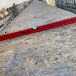 Czerwona poziomica na płycie OSB na dachu budynku w trakcie prac budowlanych, w tle zabudowa miejska.